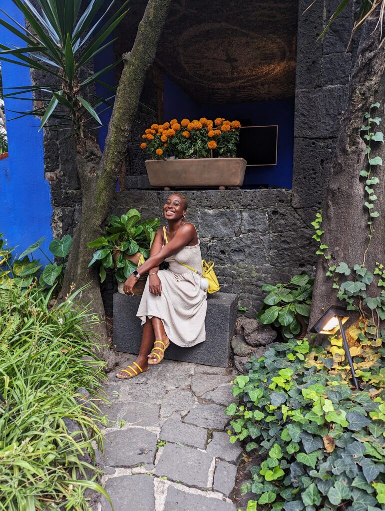 black woman with bald head sits on stone steps in a beige spaghetti strap dress and a yellow cross body bag. the stone steps face a stone path lined with green plants