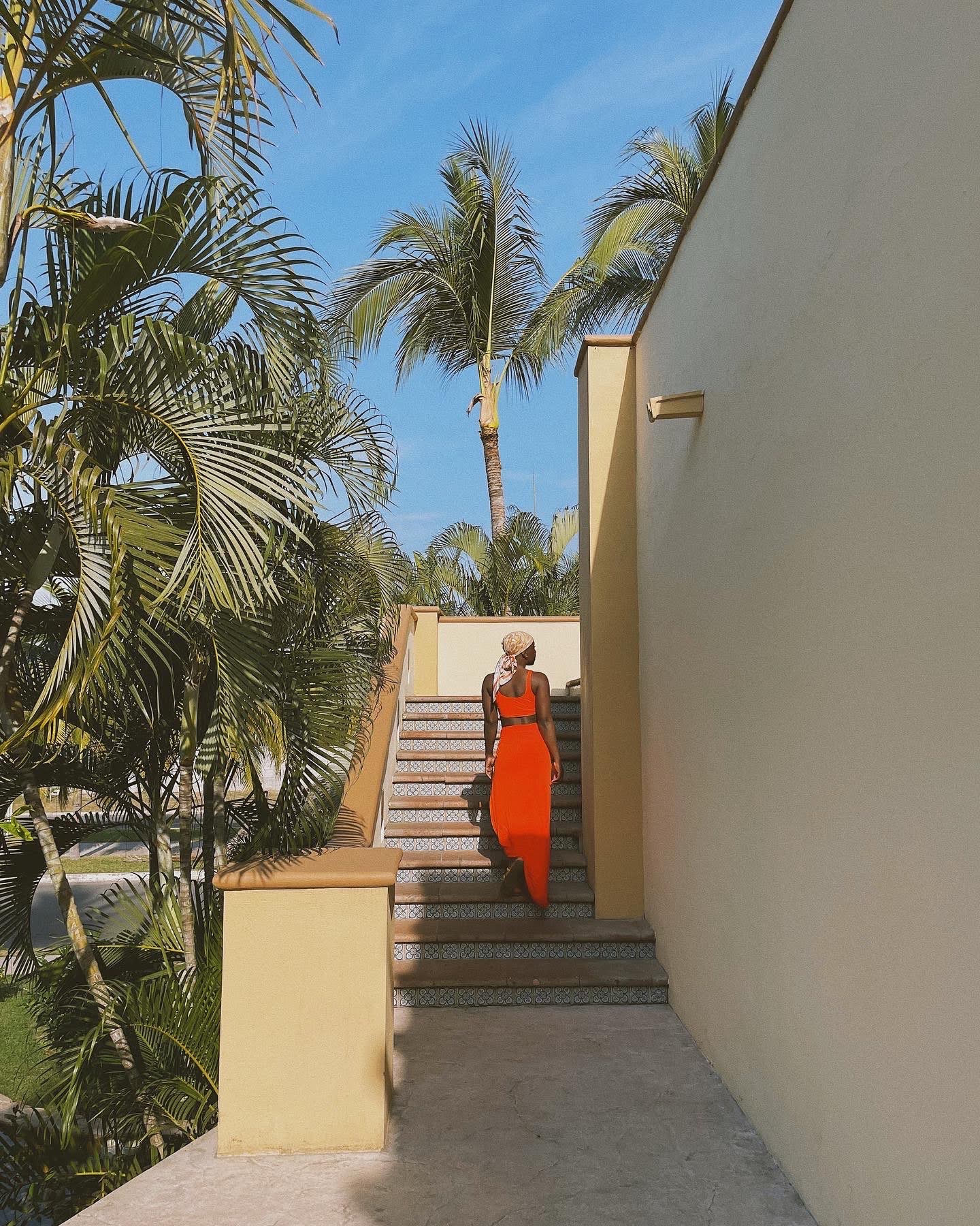 A woman in an orange two-piece swimsuit walking up staircase. Flanked by palm trees