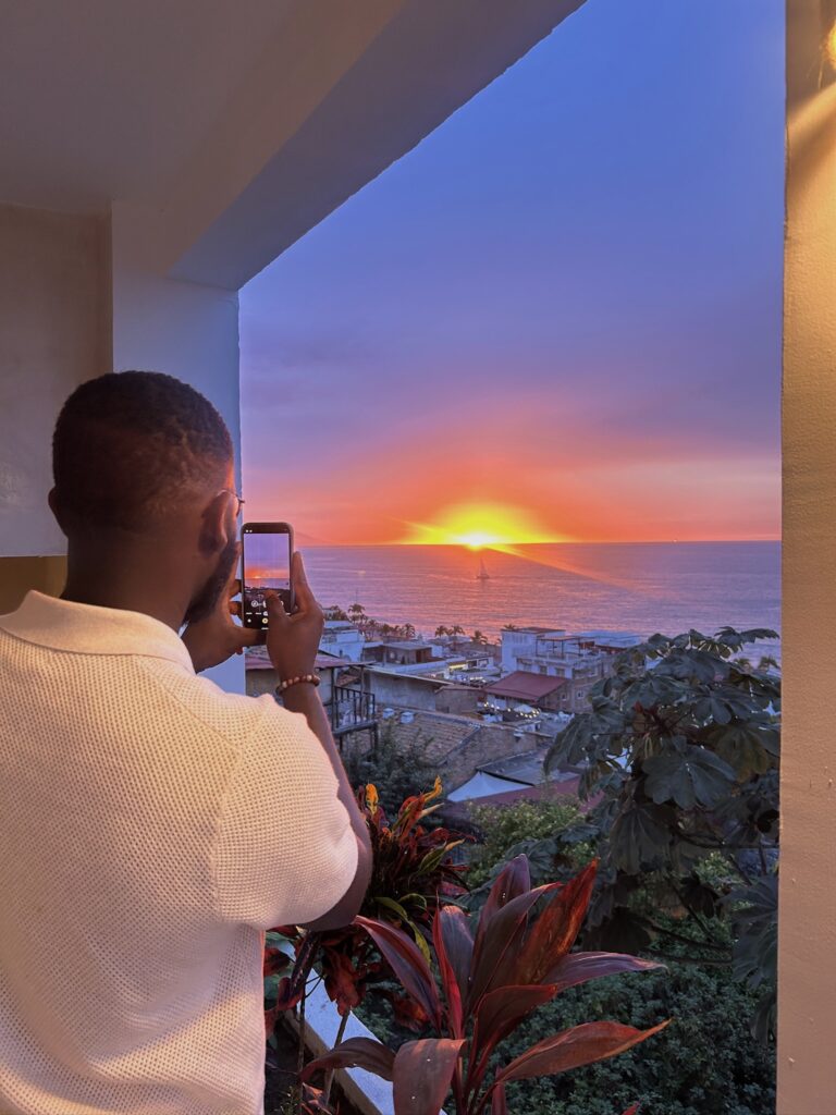 a black man holds his phone below eye level to take a picture of the sky at sunset. the sky is purple surrounding a fiery orange sun that has dipped low on the horizon