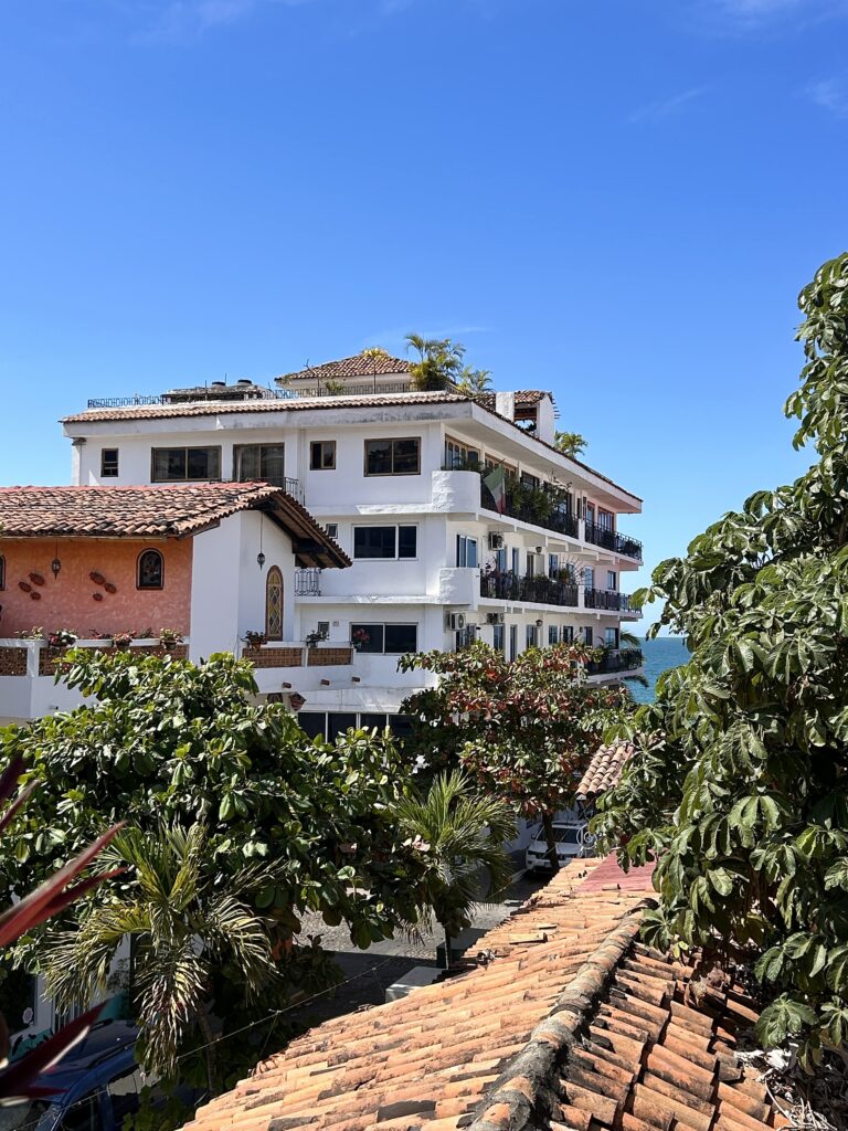A 3 story whitewashed building with balconies facing the sea, green trees fill the foreground