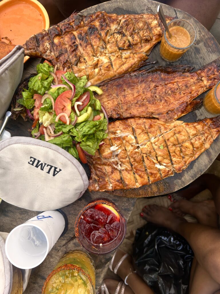a platter of grilled whole fish sits next to a bed of fresh green vegetables and two cocktails - one is  purply colored and the other is mango yellow