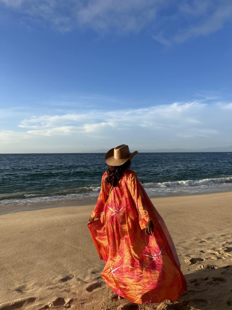a woman wearing a cowboy hat and a long orange and pink kimono, walks on the beach.