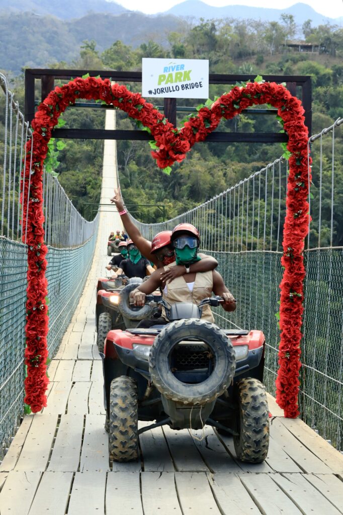 A black couple on an ATV, underneath a heart wreath made of red flowers on the Jorullo bridge in Puerto Vallarta