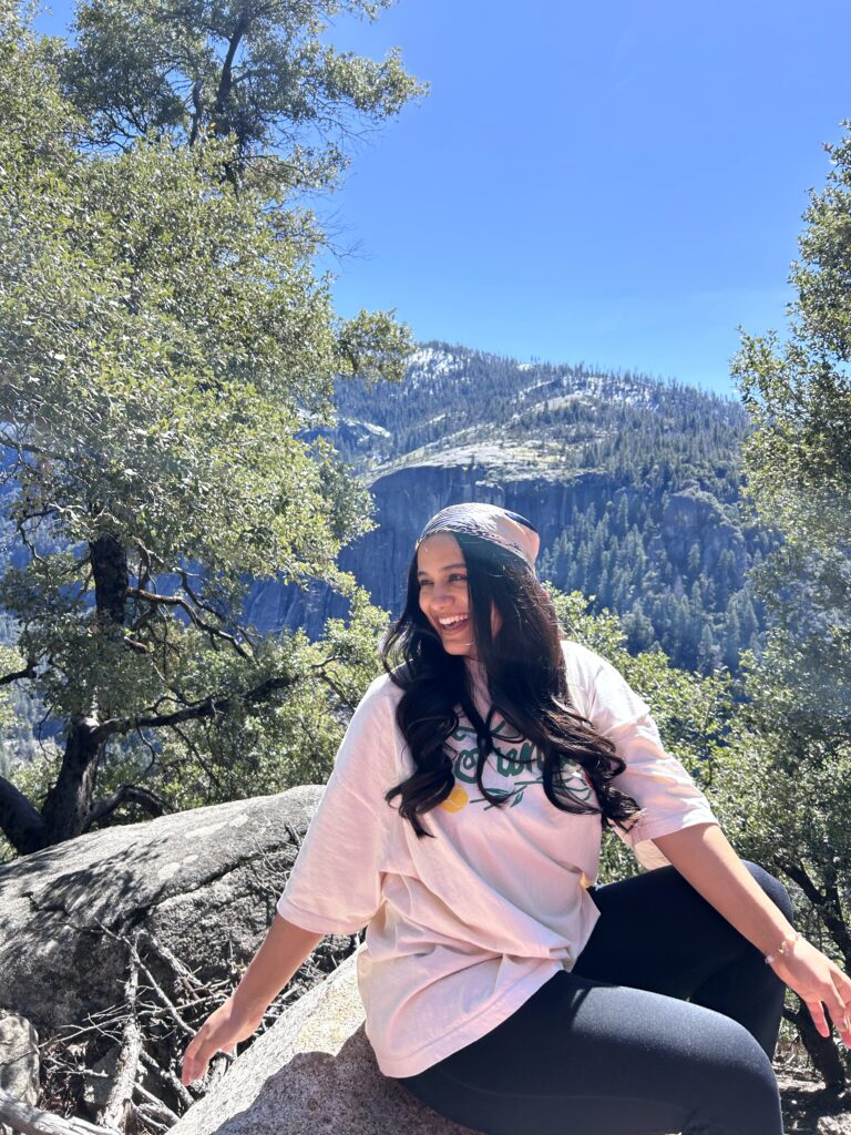 a woman with long black wavy hair covered with a scarf sits on a rock with her head turned to the left as she poses for a photo