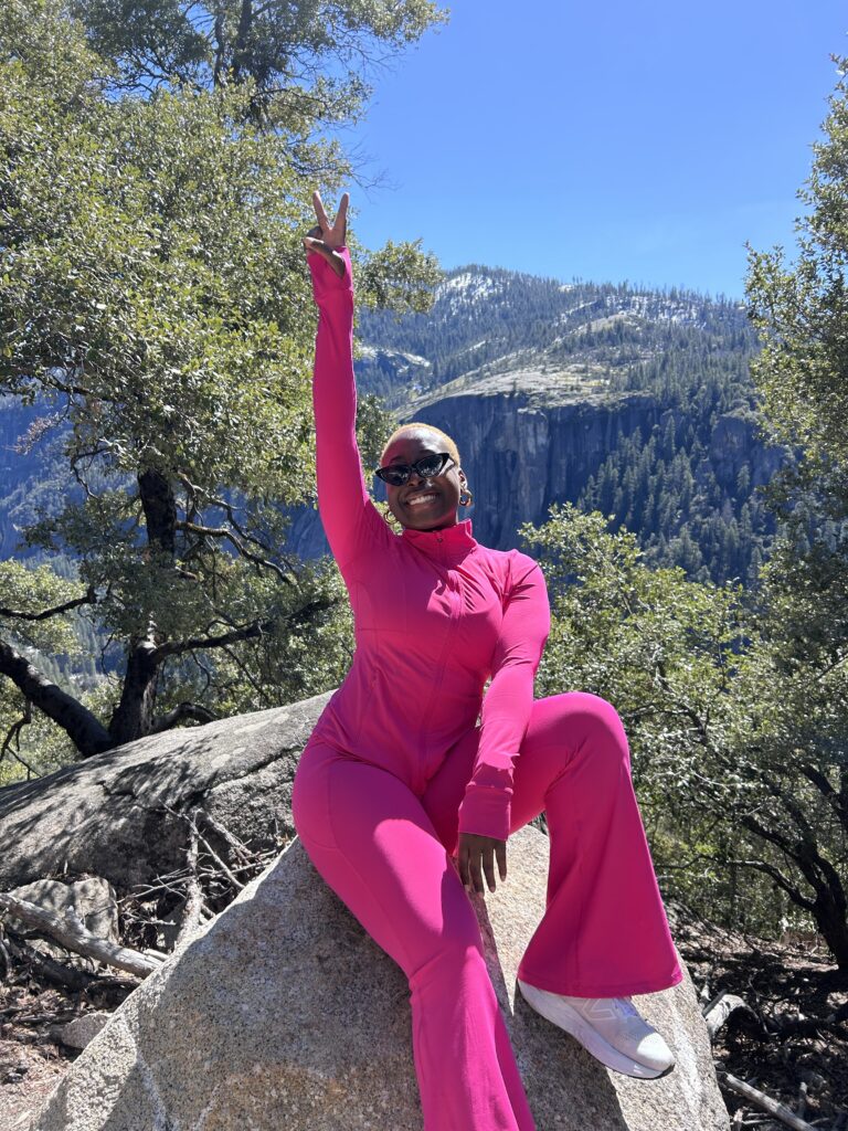 a Black woman with short blonde afro sits on a rock in an all pink athletic set smiling towards the camera.