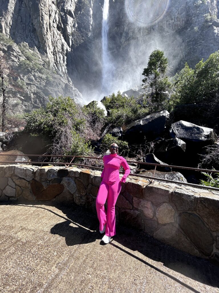 a black woman in a short blonde fro poses infront of the bridal fall cascade - a misty waterfall flowing down a gray rock face.
