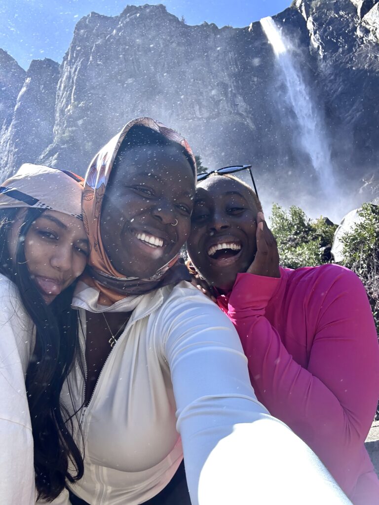 three women smile with their teeth. one has a tongue sticking out. the misty white cascade of bridal veil fall is in the background 