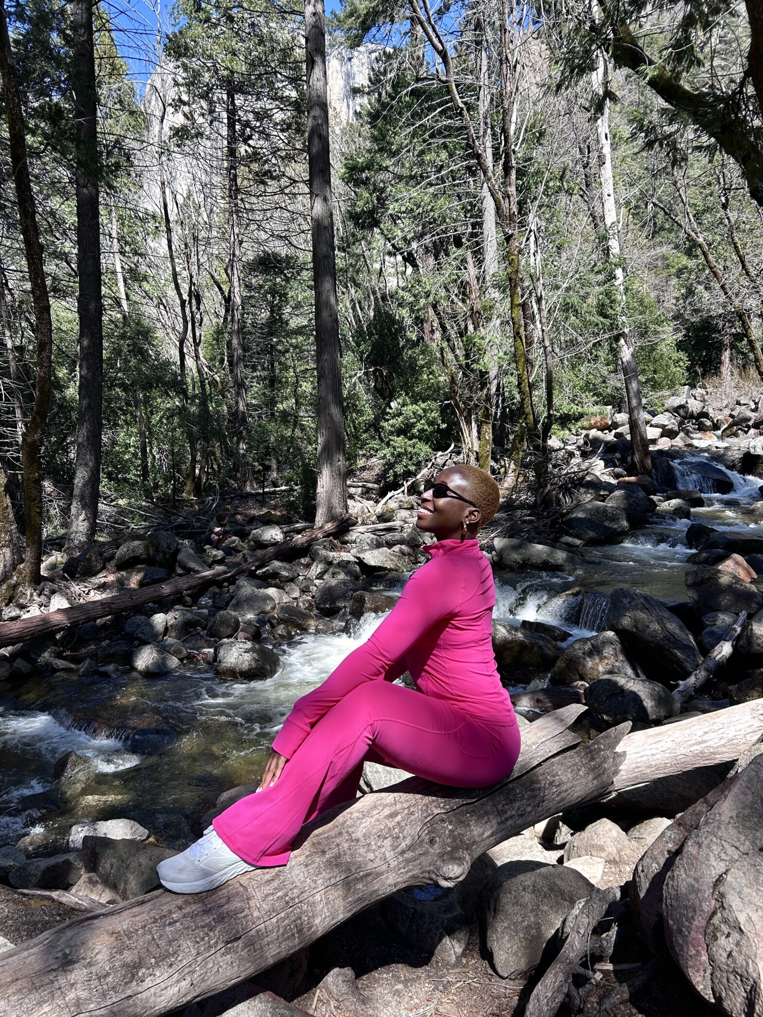 A black woman with a short blond afro sits on a wooden log thats created a natural bridge across a ravine