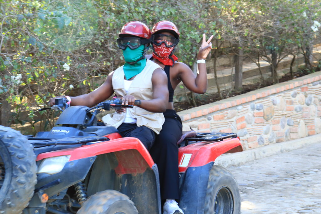 a Black couple riding on a red and black 4 wheel ATV. there is a rock river and trees in the background. the woman makes a peace sign with her fingers