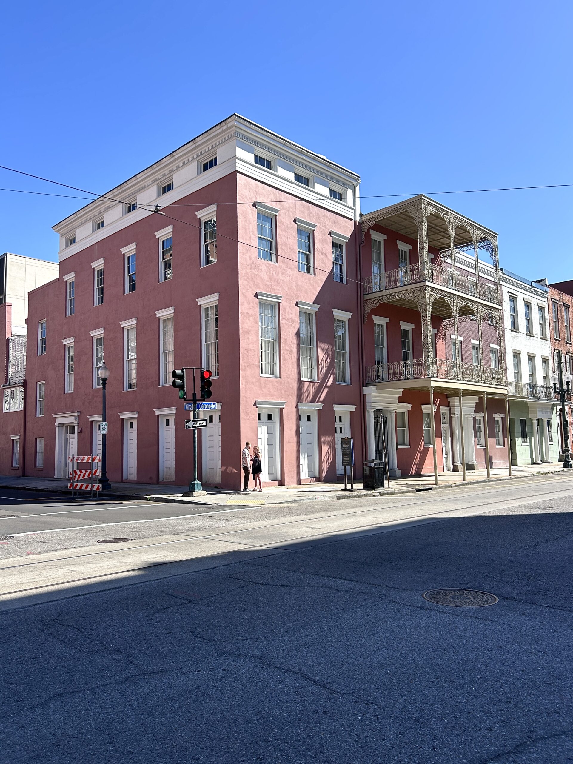 A striking pink corner building with tall white-trimmed windows and ornate wrought iron balconies sits beneath a clear blue sky in New Orleans. Two women walk along the sidewalk at the intersection of Royal and Governor Nicholls Streets, while soft shadows stretch across the pavement.