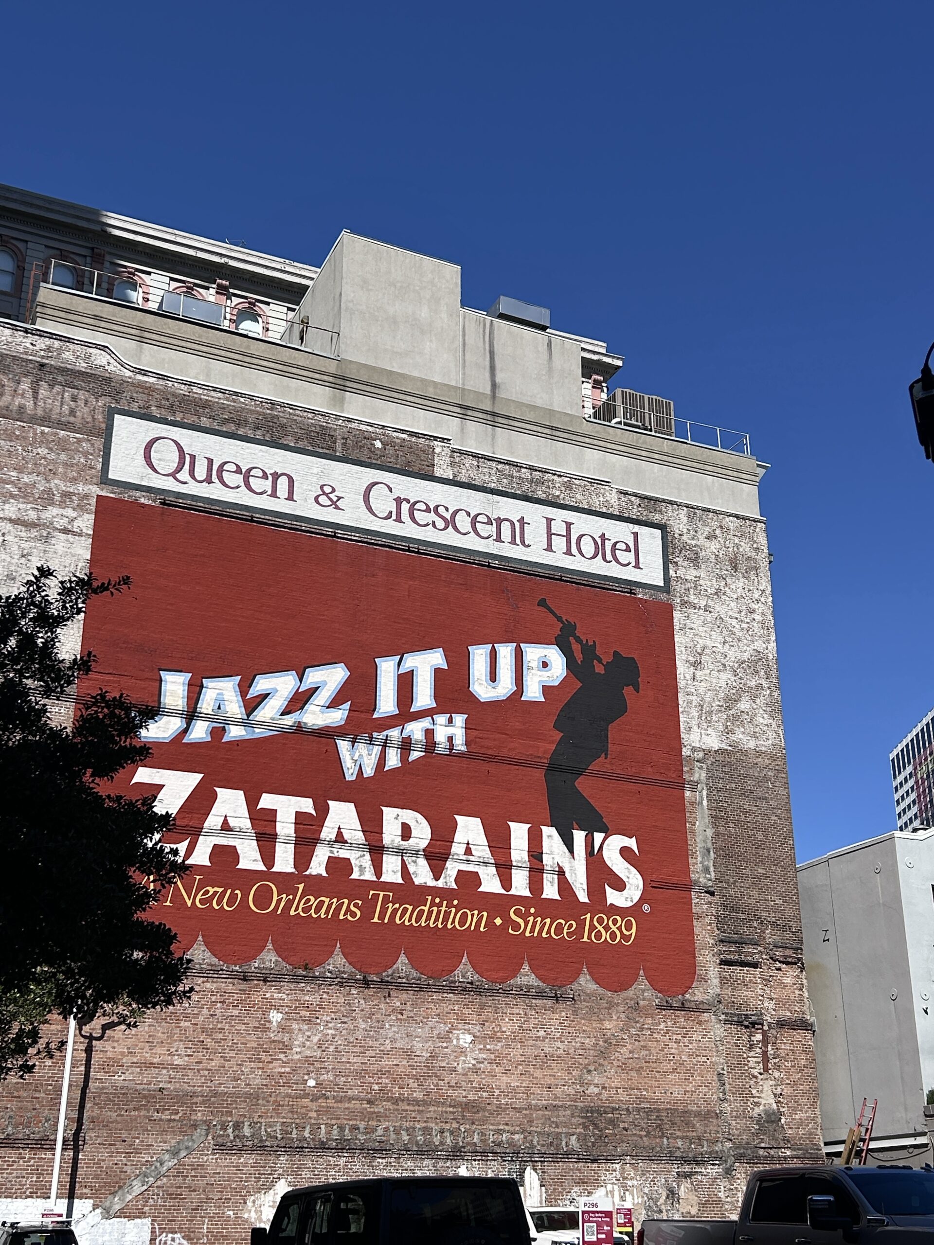 A bold red mural on the side of a brick building reads “Jazz It Up with Zatarain’s – A New Orleans Tradition Since 1889” beneath the Queen & Crescent Hotel sign. A jazz musician silhouette adds flair to the vintage-style advertisement.