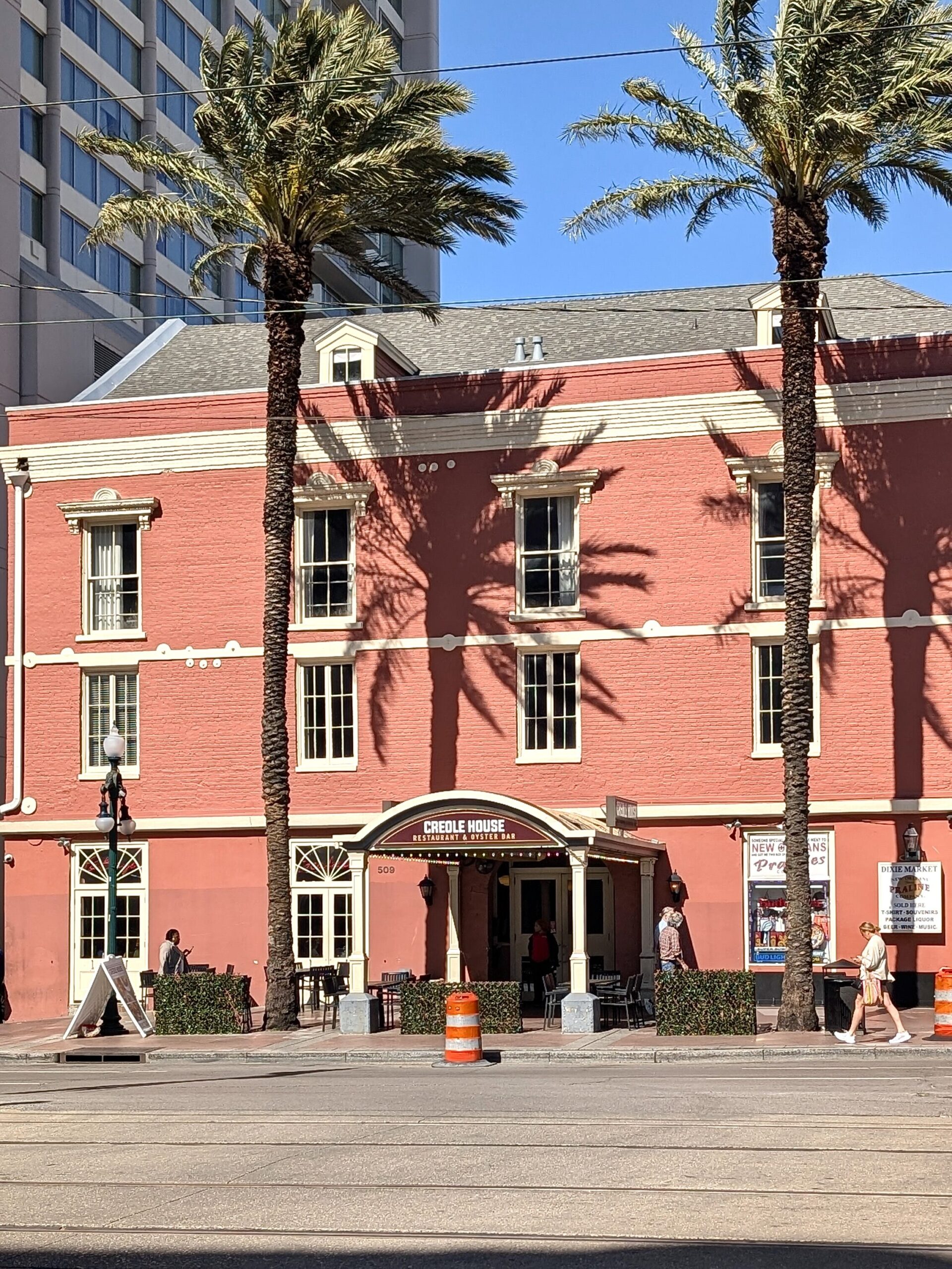 The Creole House Restaurant & Oyster Bar stands out with its salmon-colored brick exterior and white trim. Palm tree shadows stretch across the building, and a few pedestrians walk along the sunlit street in front.