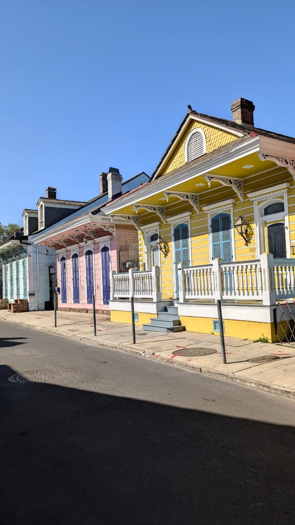 A row of vibrant New Orleans shotgun houses, painted in pastel shades of yellow, pink, and turquoise, lines a quiet residential street under a cloudless blue sky. Decorative trim and shutters add charm to the bright façades.