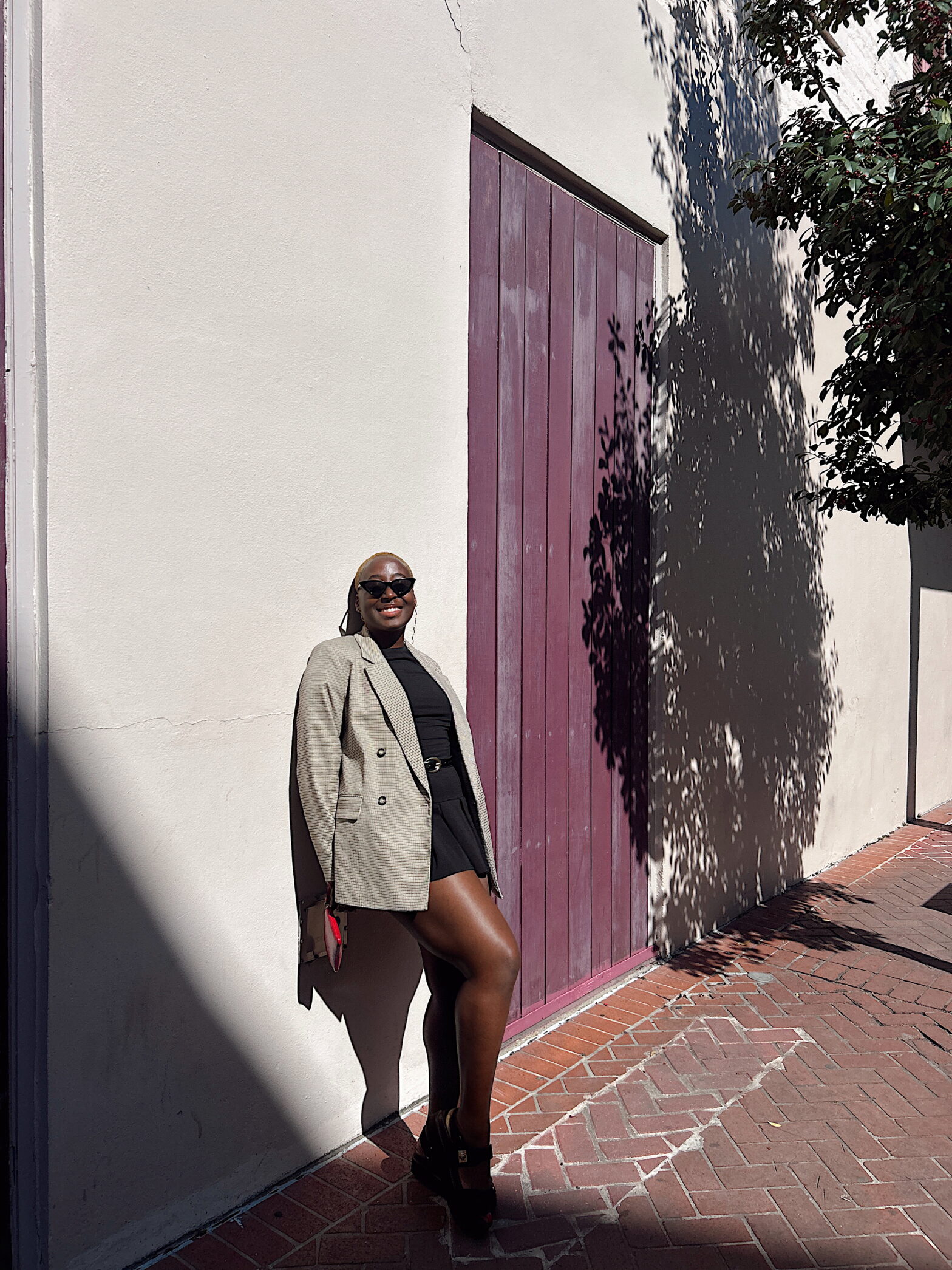 A smiling woman leans against a pale wall between plum-colored wooden doors. Dressed in a black outfit with an oversized blazer, she stands in warm sunlight while tree shadows dapple the surface beside her.