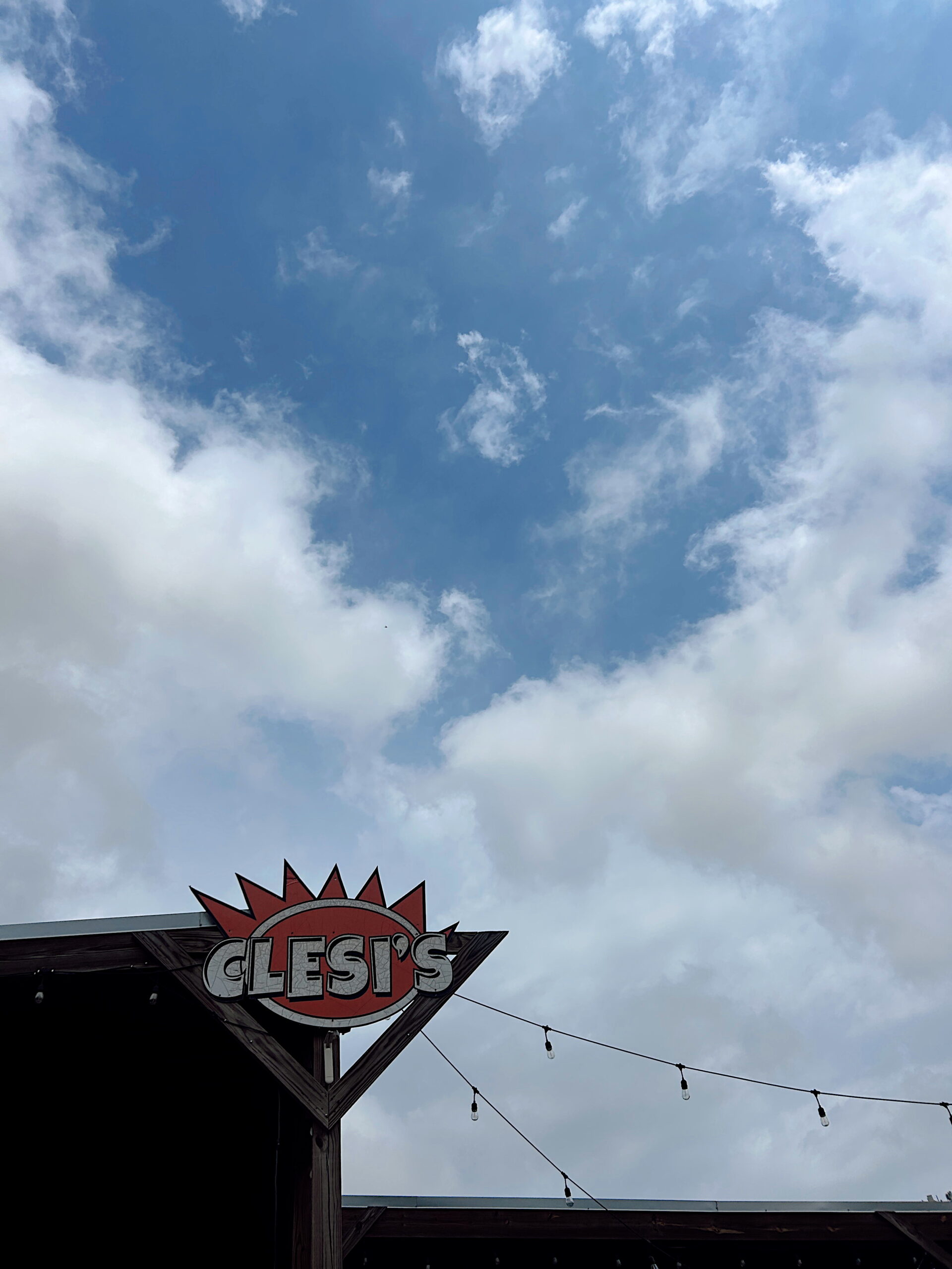 A weathered wooden sign reading “Clesi’s” is mounted above a patio adorned with string lights. The sign stands out against a partly cloudy blue sky.
