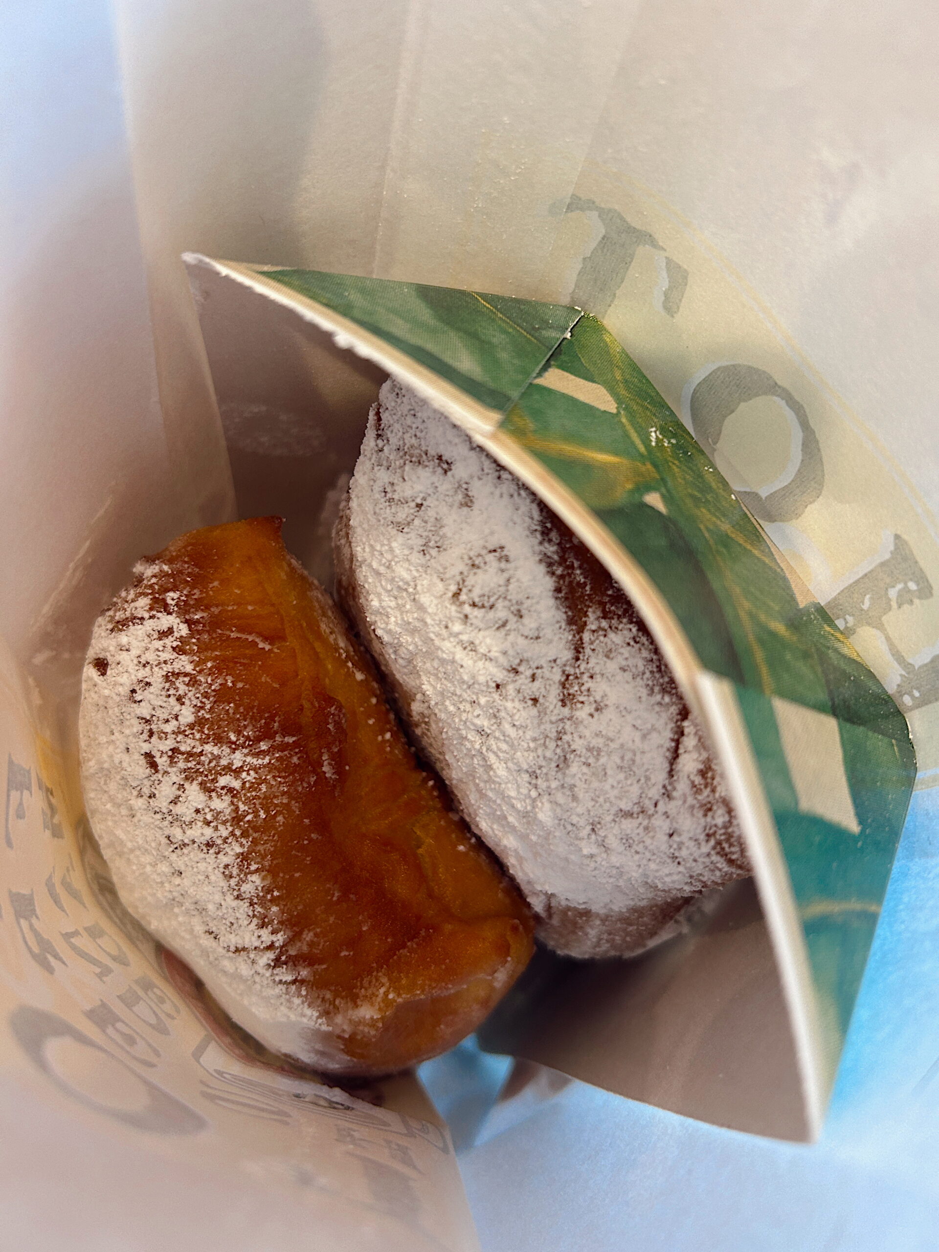 Two golden-brown beignets generously coated in powdered sugar peek out from a branded paper bag, ready to be enjoyed.