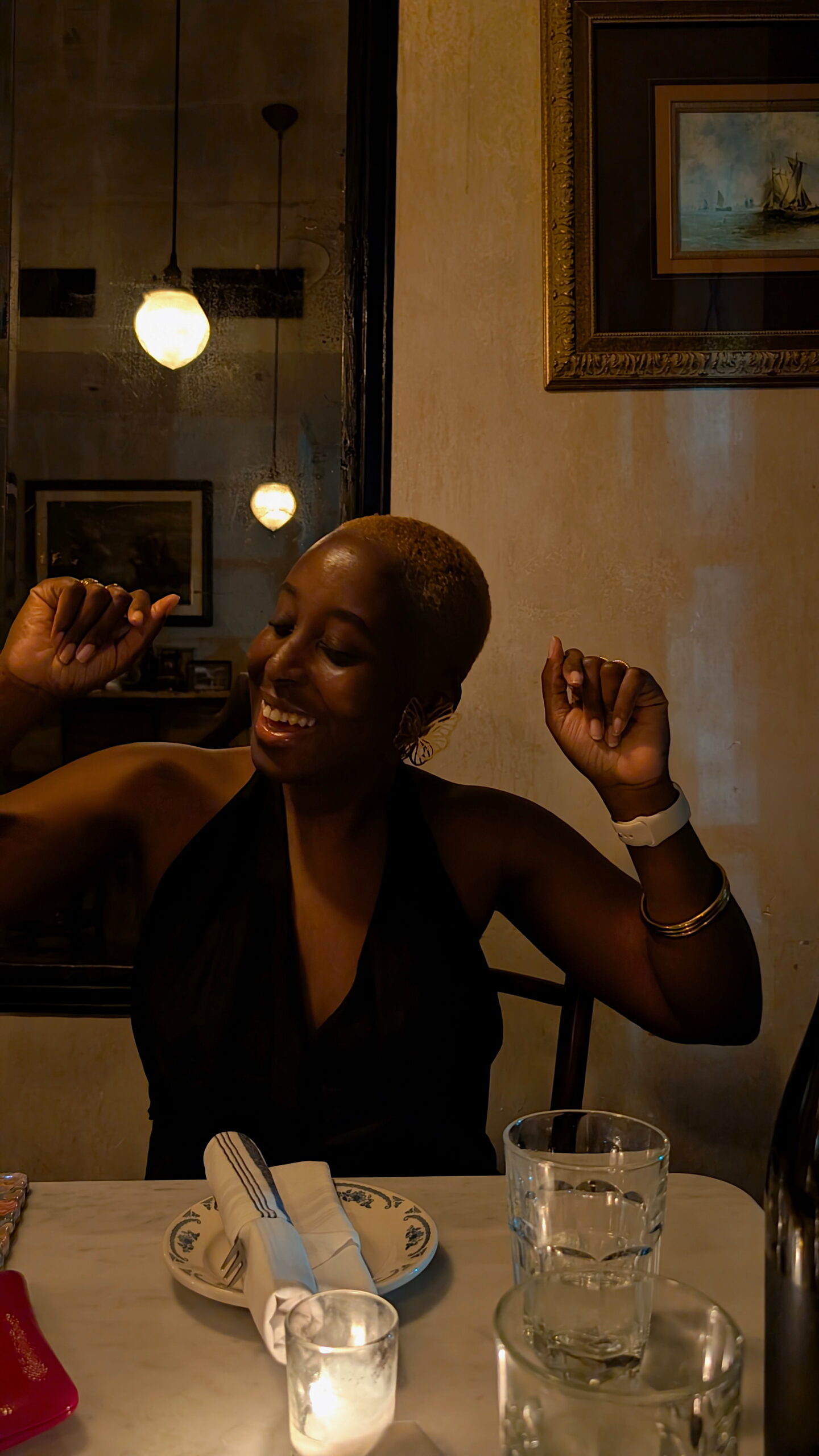 A woman with short blond hair sits at a candlelit dinner table, joyfully raising her arms mid-dance and laughter. She’s dressed in a halter top, surrounded by softly glowing pendant lights and vintage-style decor, creating a warm and celebratory mood.