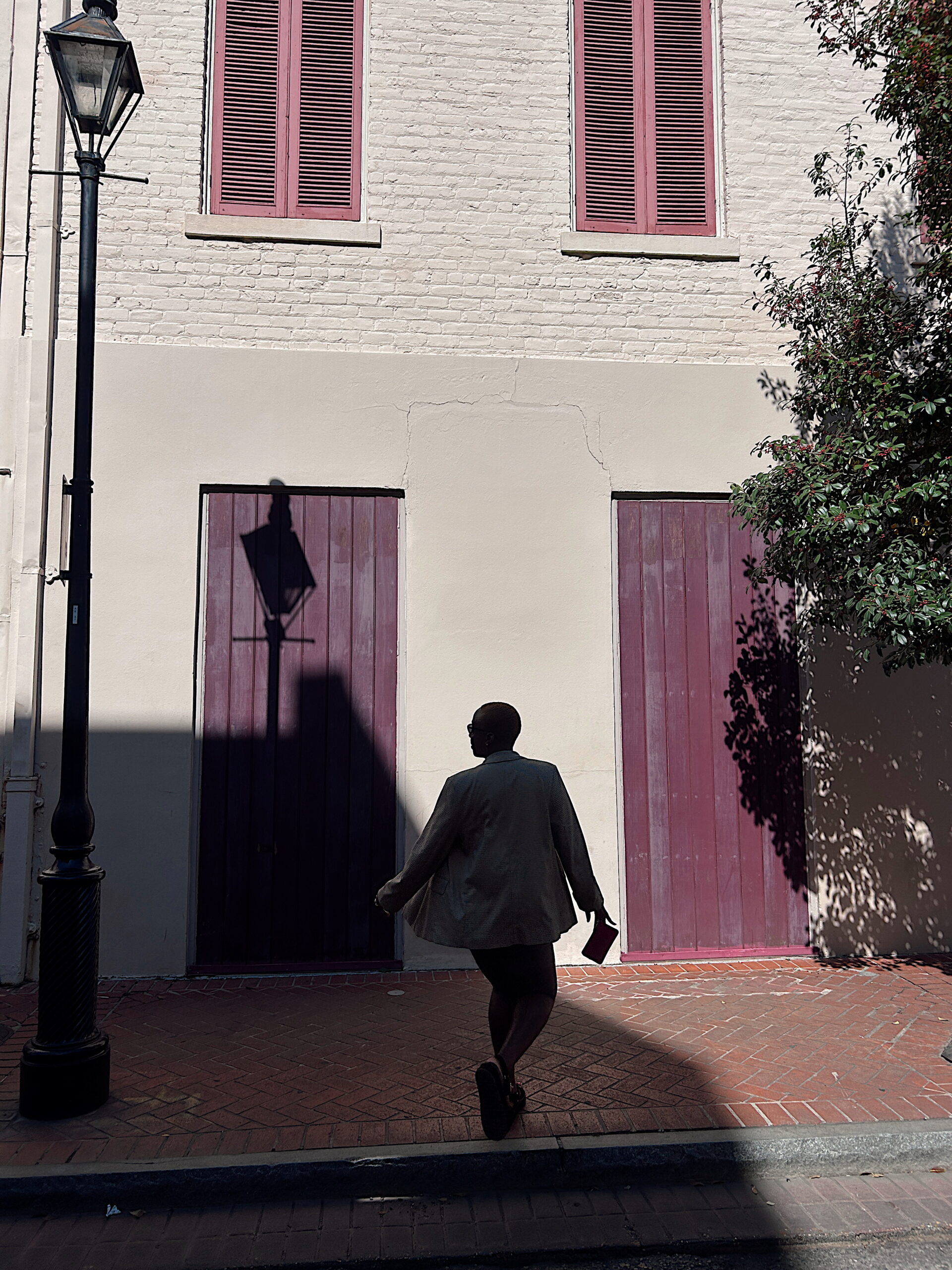 A person in a blazer and sandals walks toward a cream-colored building with deep plum doors and shutters. The midday sun casts sharp shadows of a streetlamp and tree branches across the brick sidewalk and wall.