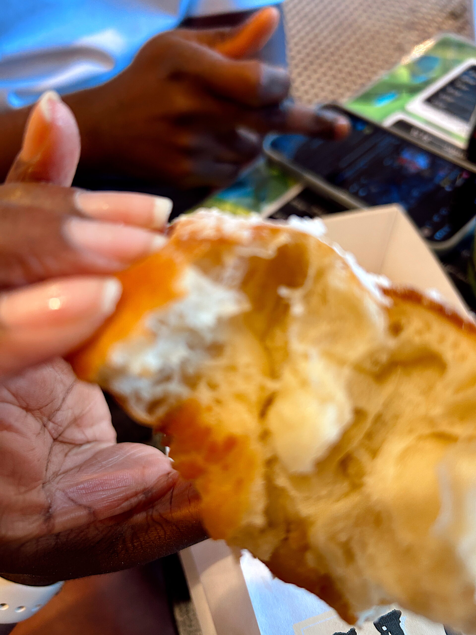 A hand holds a partially eaten beignet, its airy interior and light dusting of powdered sugar visible. In the background, another hand gestures over a phone, suggesting a casual café setting.