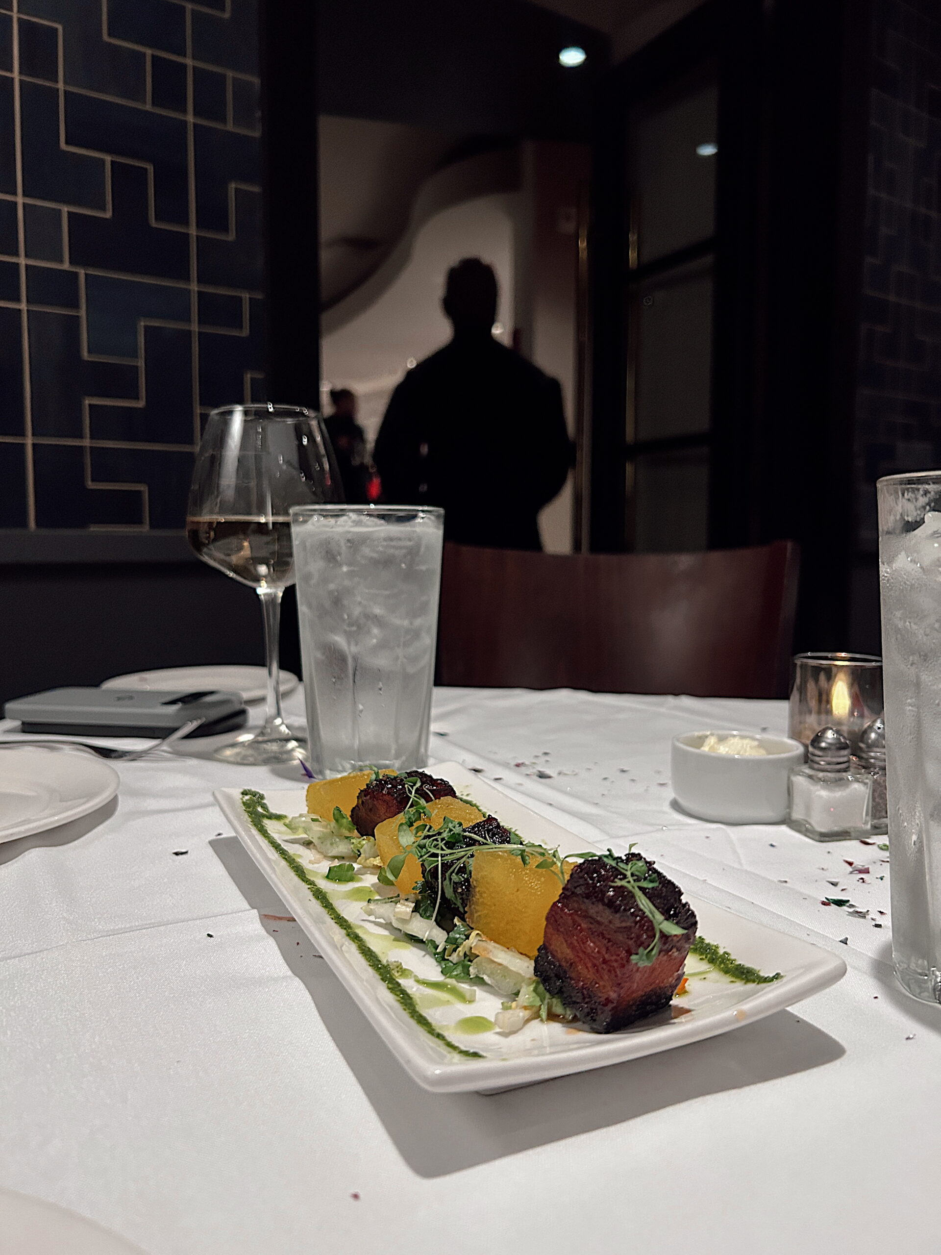 A plated appetizer featuring seared cubes of meat, vibrant yellow beets, and microgreens is artfully arranged on a rectangular dish, garnished with green sauce and finely chopped vegetables. The table is set with a wine glass, water, and a small candle, all resting on a white tablecloth scattered with festive confetti. In the background, a silhouetted figure stands in a softly lit restaurant entryway.