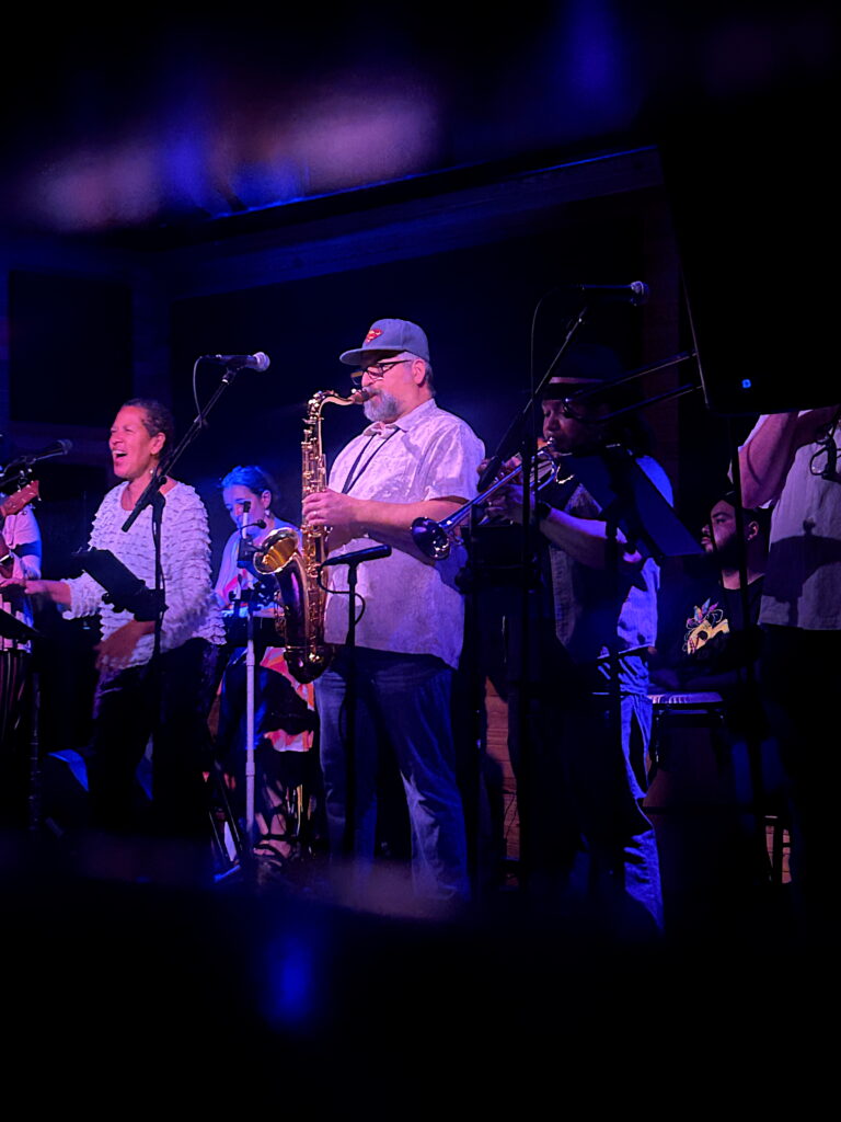 A live band performs on stage under dramatic purple-blue lighting. A saxophonist in a cap plays center stage, flanked by a smiling female vocalist and fellow brass players. The scene is vibrant and full of energy, capturing the joy of live music.
