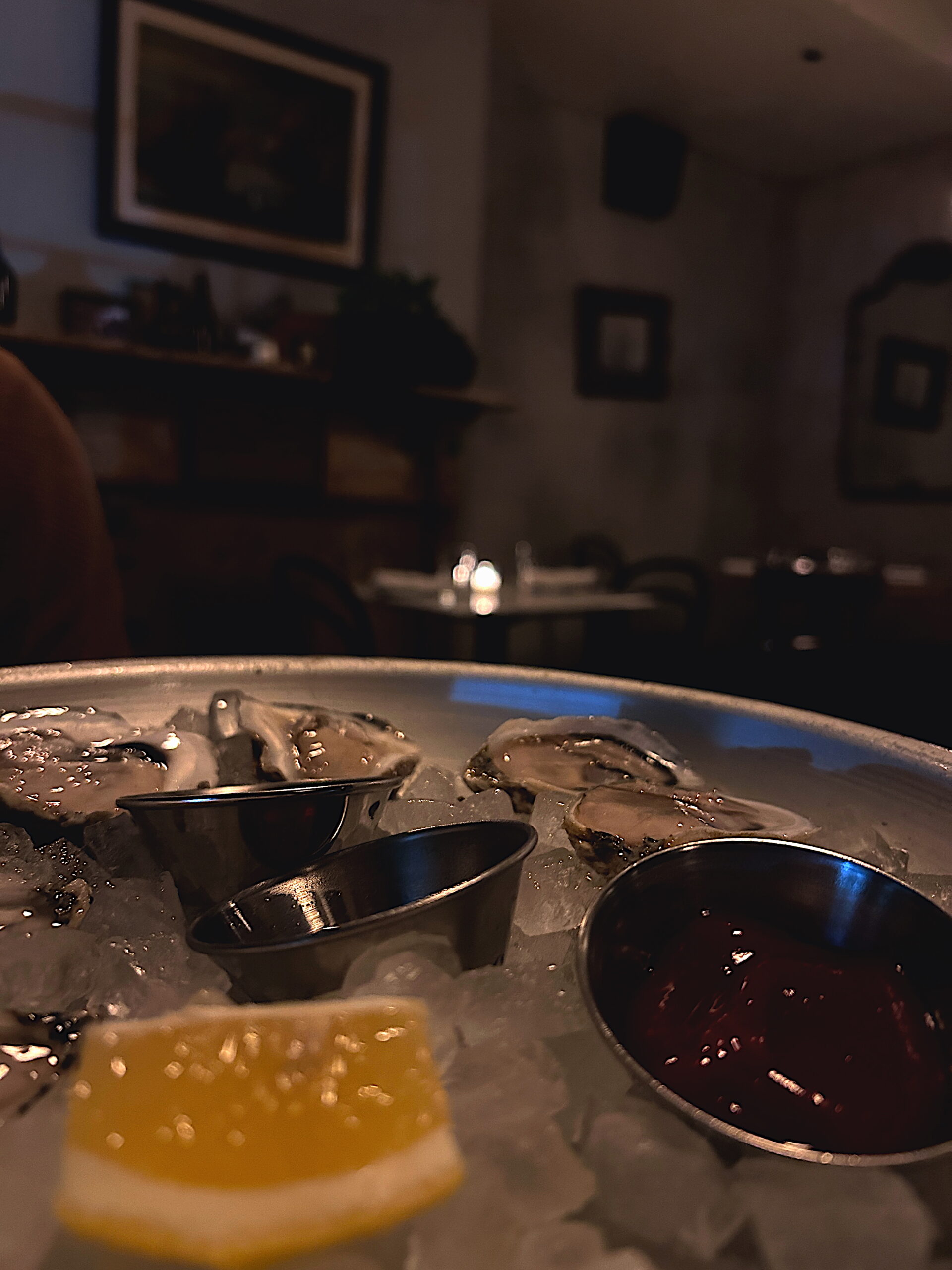 A close-up shot of freshly shucked oysters on a bed of ice, served with lemon wedges and metal sauce cups. The scene is warmly lit, capturing the cozy ambiance of a dimly lit restaurant.