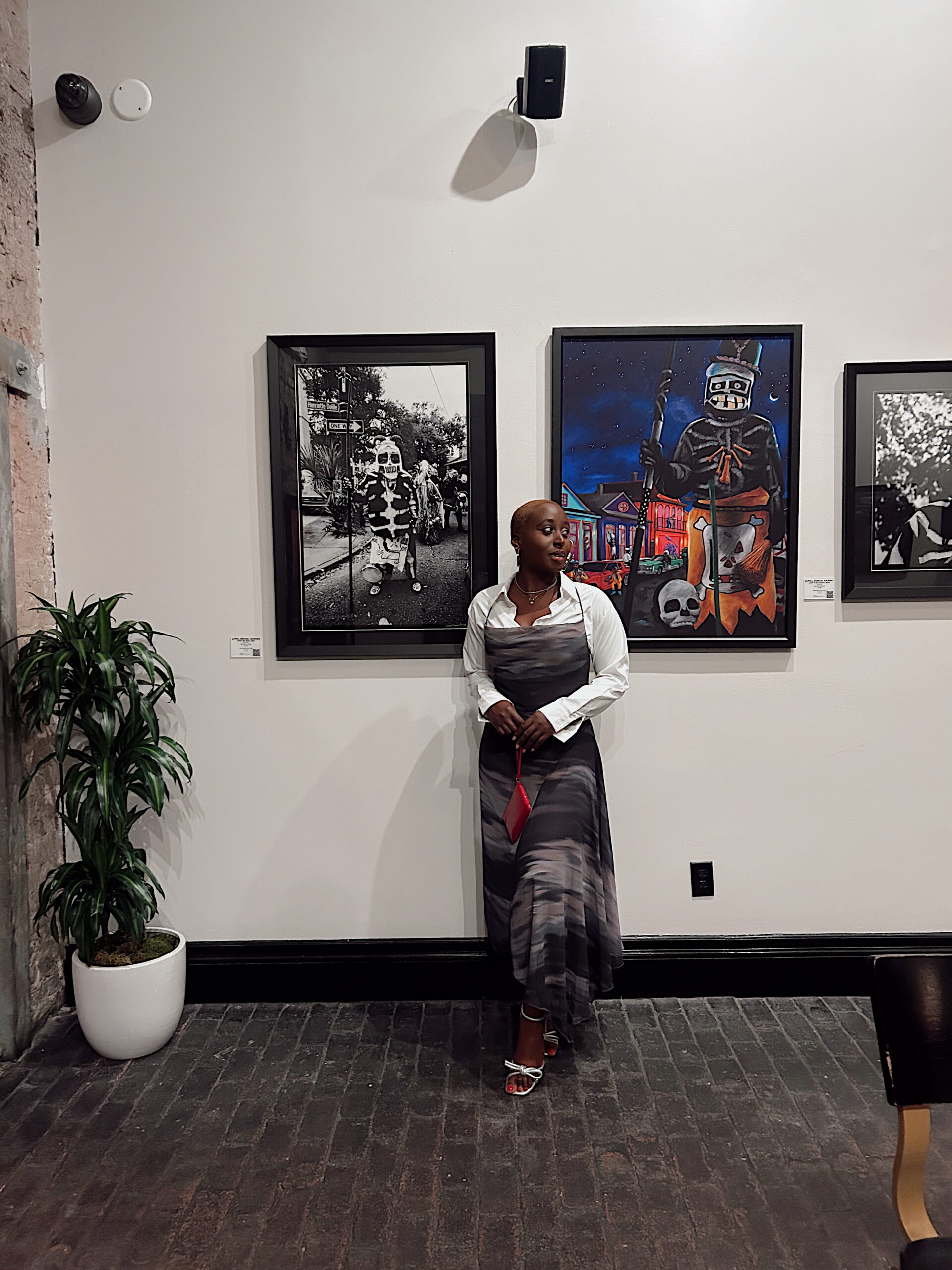 A woman poses in a long, tie-dye-style dress and white heels in an art gallery, framed between two pieces of art—one black-and-white photograph and one vibrant painting. A red clutch adds a pop of color to her polished look.