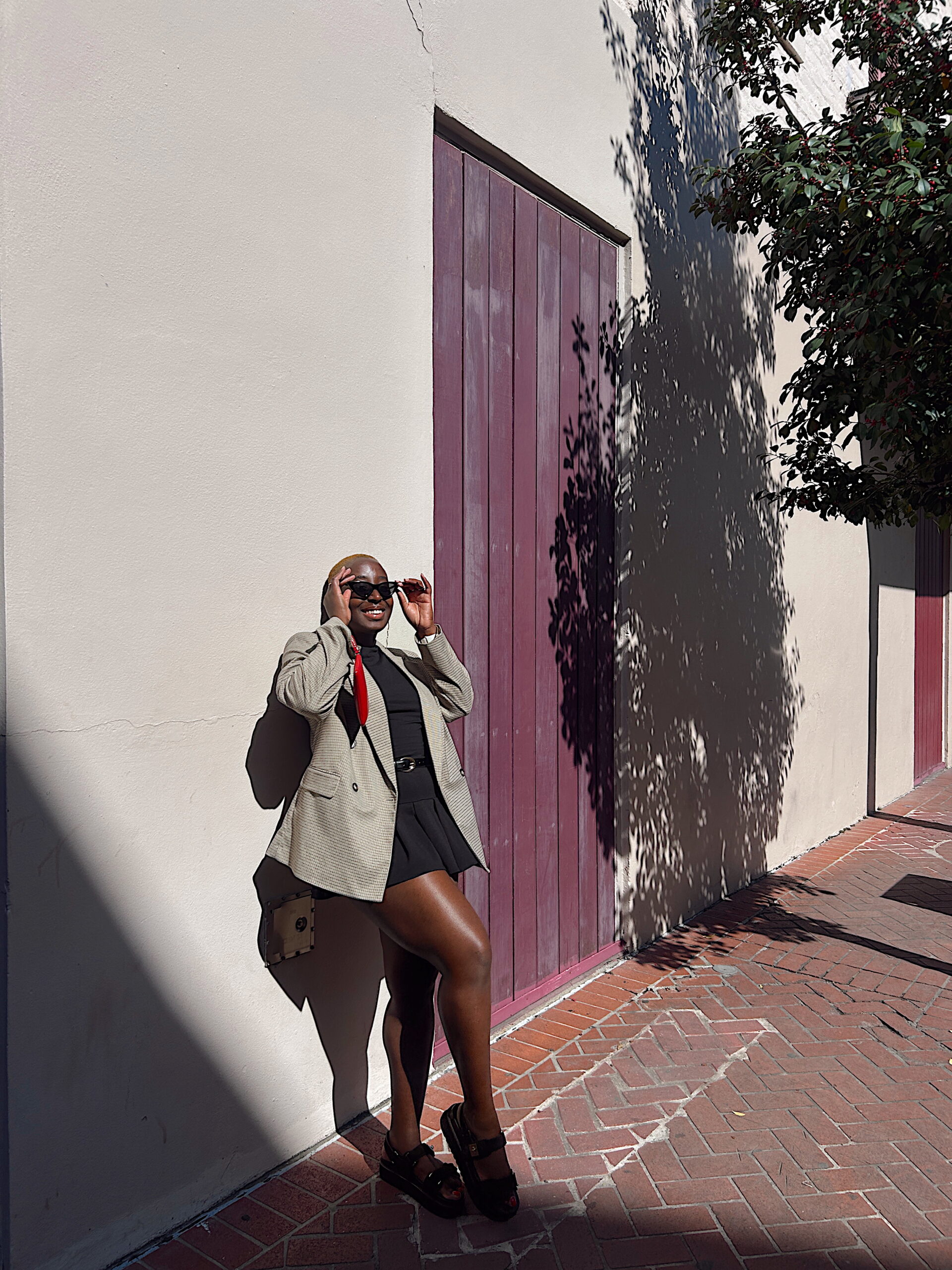 A smiling woman leans against a pale wall between plum-colored wooden doors. Dressed in a black outfit with an oversized blazer, she stands in warm sunlight while tree shadows dapple the surface beside her.