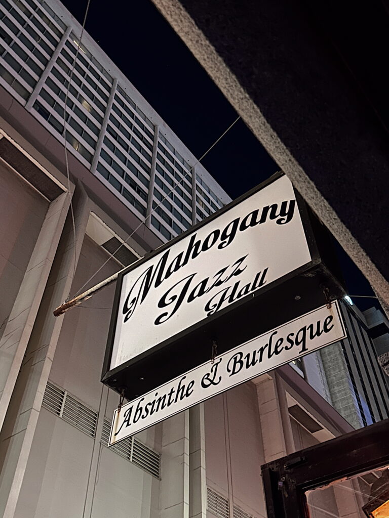 A nighttime street view featuring the Mahogany Jazz Hall sign, which reads “Mahogany Jazz Hall – Absinthe & Burlesque.” The sign hangs against the backdrop of a tall, modern building, contrasting the club’s vintage allure with the city’s urban architecture.