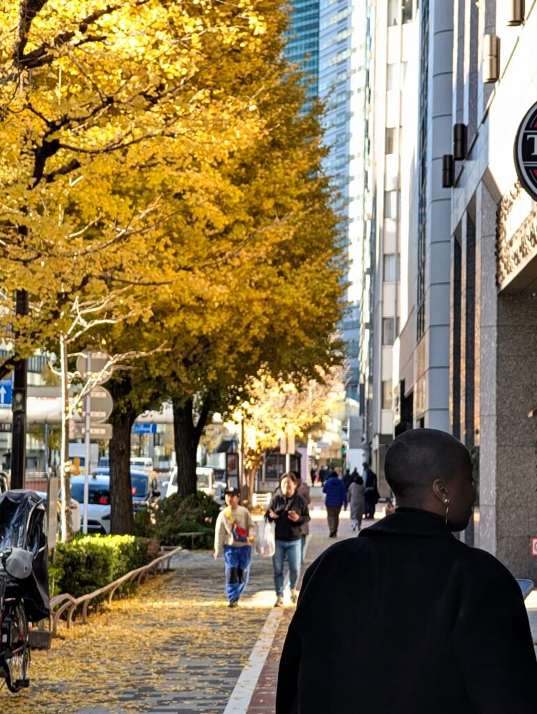 A black woman walks on the street wearing a black coat. her hair is bald. there is a row of yellow leaved trees to her left.
